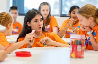 image of tutor and children in conversation wearing orange shirts sitting around white tables 