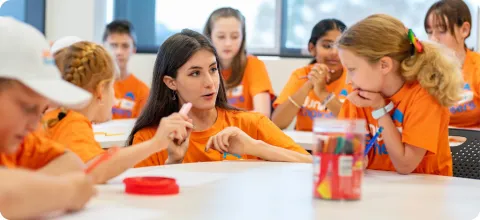 image of tutor and children in conversation wearing orange shirts sitting around white tables 